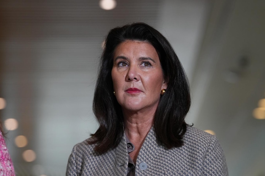 A dark-haired woman in a houndstooth jacket stands in a corridor.