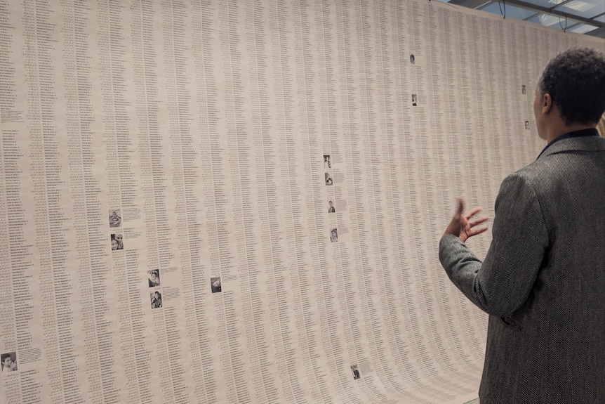 A man in a grey jacket gestures with his hands as he looks at a list of thousands of names