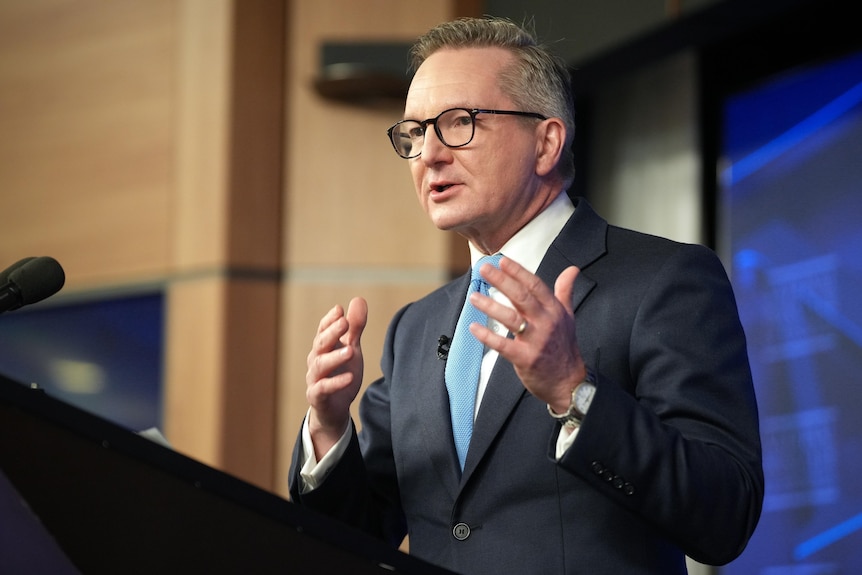 Chris Bowen, wearing a suit with a blue tie and glasses, speaks at a podium with a microphone.