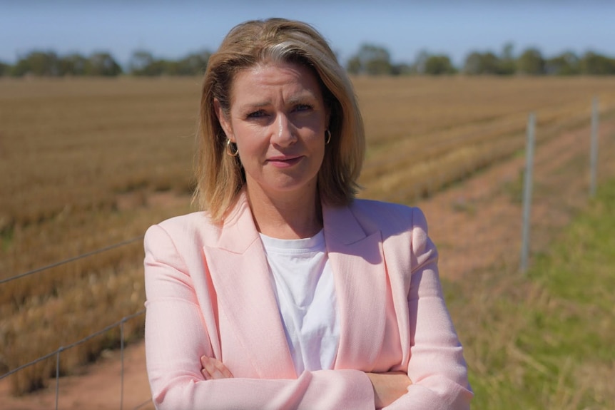 A woman wearing a pink blazer and white tshirt stands in front of farmland