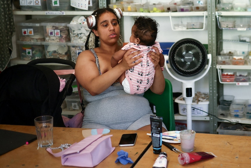 A woman holds her baby as she sits near a fan.