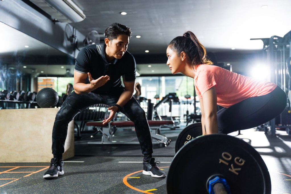 A male personal trainer coaches a woman on lifting weights in a gym.