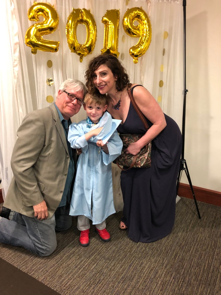 A man with white hair and dark glasses, wearing gray jeans and a tan jacket, kneels left. A little boy in a blue graduation gown, holding his cap, stands center. Crouched next to him at right is a dark-haried woman in a sleeveless black dress, holding a brown patterned purse. Behind them is a white curtain and gold balloons spelling out 2019.