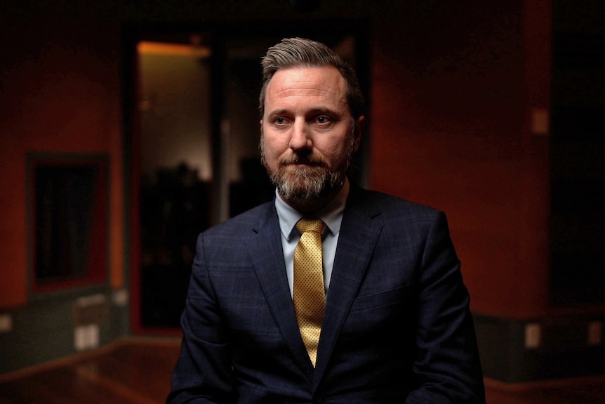 A man wearing a suit and tie sits in a darkened room. He is looking ahead with a neutral expression.