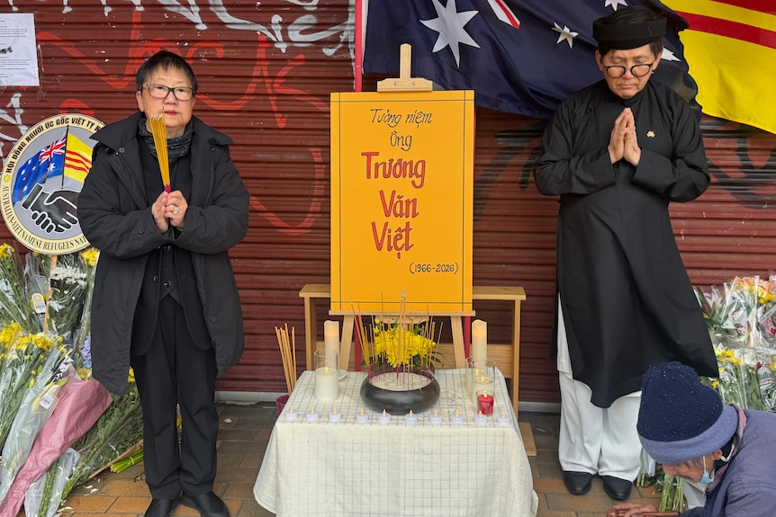 A man and woman dressed in black flank a sign that says Truong Van Viet with incense and flowers.