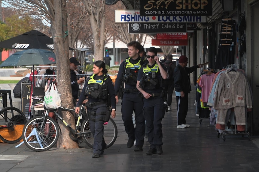 Three police officers in navy uniforms and bright yellow vests walk along a suburban shopping street on a sunny day.