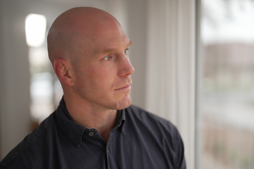 a bald man wearing a black collared shirt looking out of a window with a serious expression