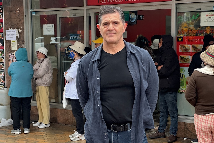 A dark haired man in an opened navy collared shirt with a black t-shirt underneath and jeans stands near people outside a shop.
