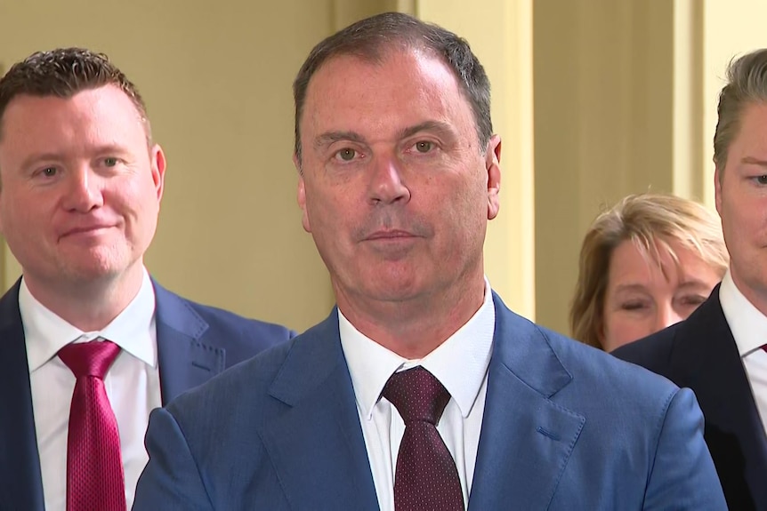 A dark haired man in a blue jacket, white shirt and dark red tie stands in front of two other men.