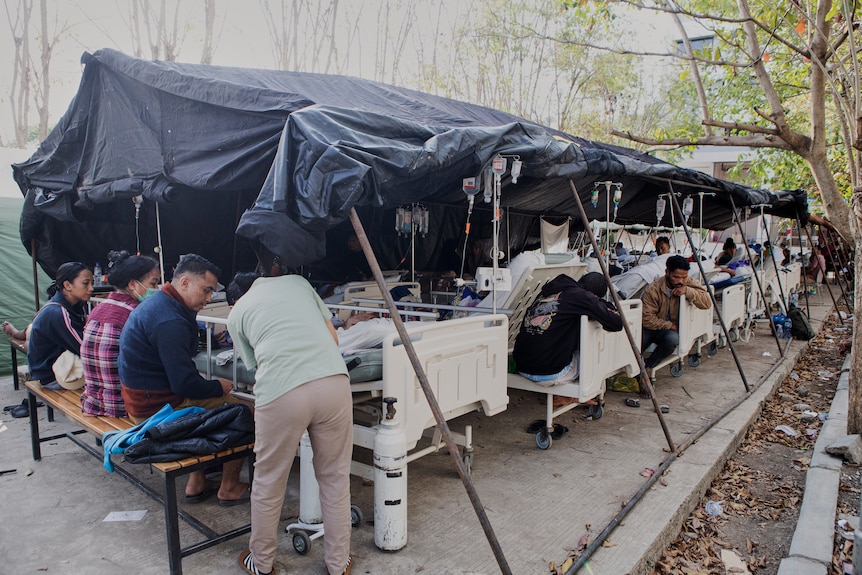 A row of hospital beds outdoors under a roof of black tarpaulin.