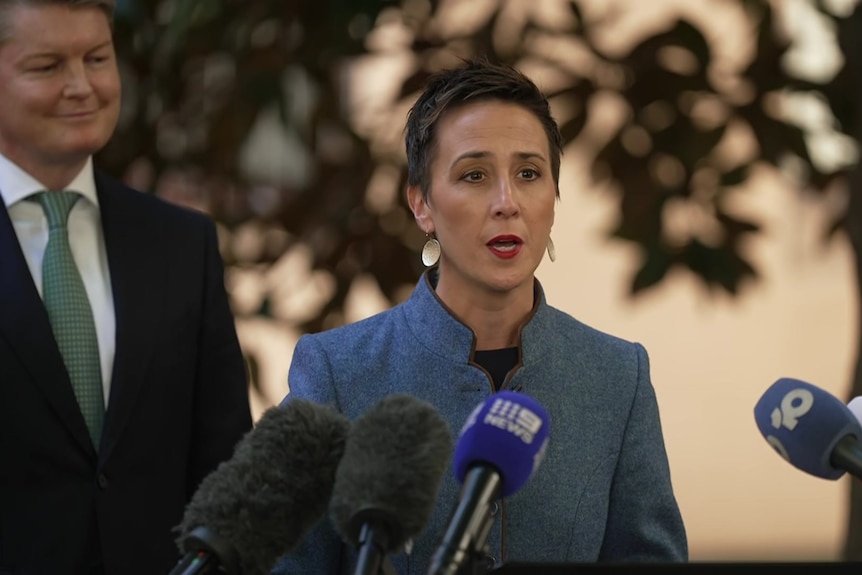 A man in a black blazer and green tie smiles looking at a woman with short dark hair in blue blazer talking into mics.