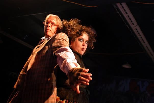 A man in a striped suit stands left, and a woman with flyaway dark hair and messy dark eye makeup stands right. They are back to back, their arms outstretched and hands touching.
