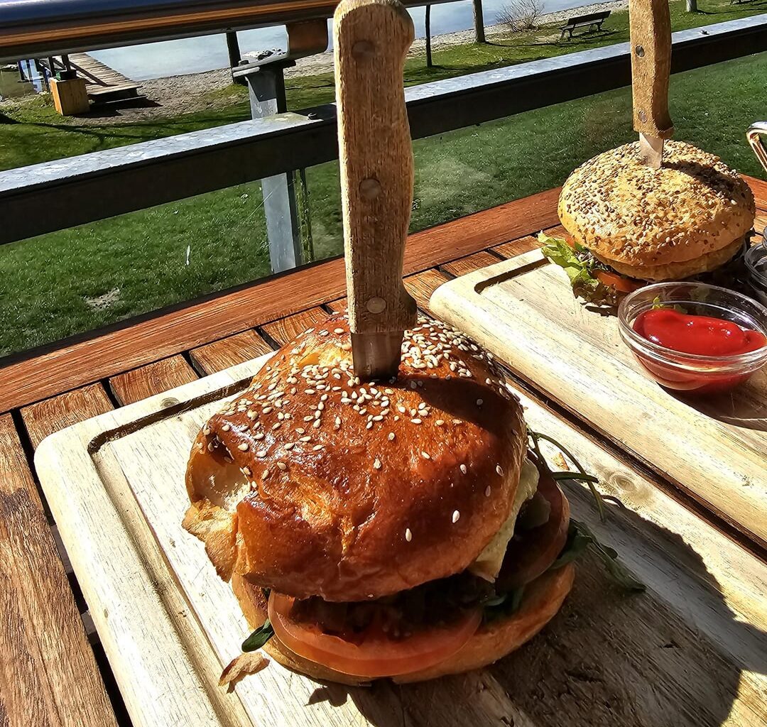 Beef Burger (minced beef, cheese, tomatoes, gherkins, melted onions, rocket) with a side salad; and a Vegan Burger (with a cauliflower patty, lettuce, tomato, cucumber, sprouts and chive cream) with fries; served at the lake near Radolfzell, Germany
