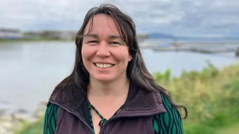 Marianne Green smiling at the camera on Rathlin Island. She has long, dark hair and is wearing a dark-coloured bodywarmer over a green and black patterned top.