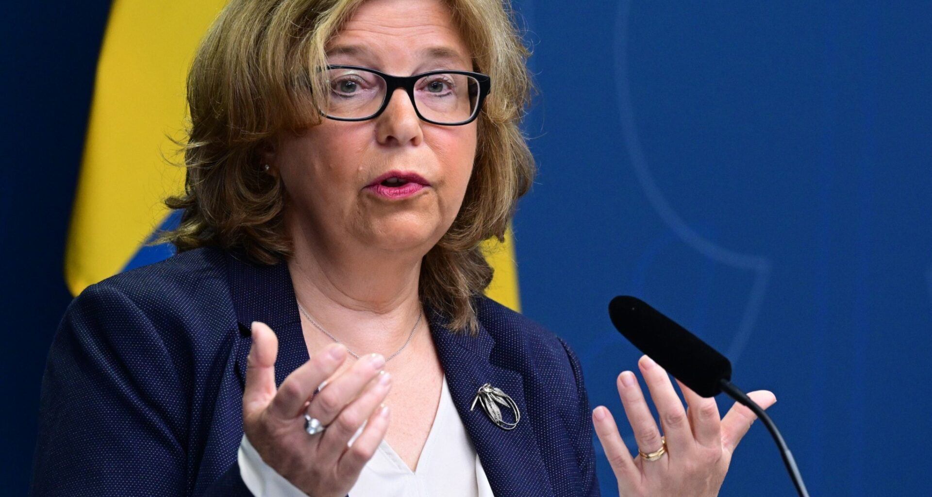 Kvinna med mellanblont hår och blå kavaj och vit blus står framför en svenska flagga.