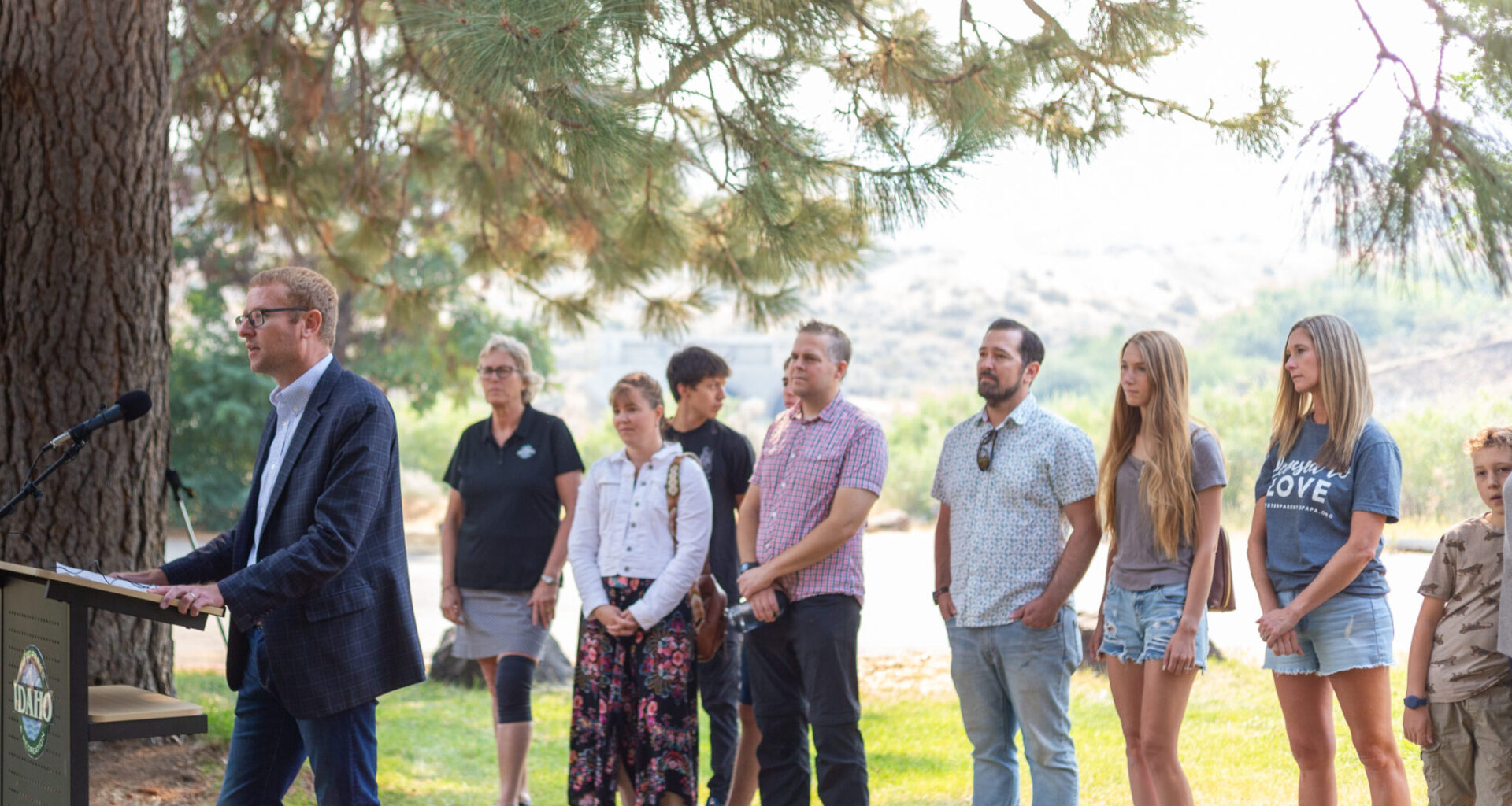 Idaho Department of Health and Welfare Director Alex Adams speaks at a podium, in front of a line of foster care families, parents and state officials