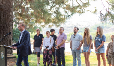 Idaho Department of Health and Welfare Director Alex Adams speaks at a podium, in front of a line of foster care families, parents and state officials