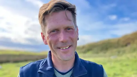 Ulf Keller looking to the camera on a sunny day on Rathlin Island. He has short dark-blond hair and is wearing a blue bodywarmer over a green jumper and navy T-shirt.