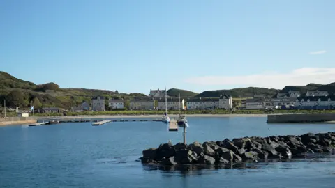 A view of Church Bay on Rathlin Island on a sunny day where a cluster of buildings line the shore around a harbour. Two small boats are docked in the bay.