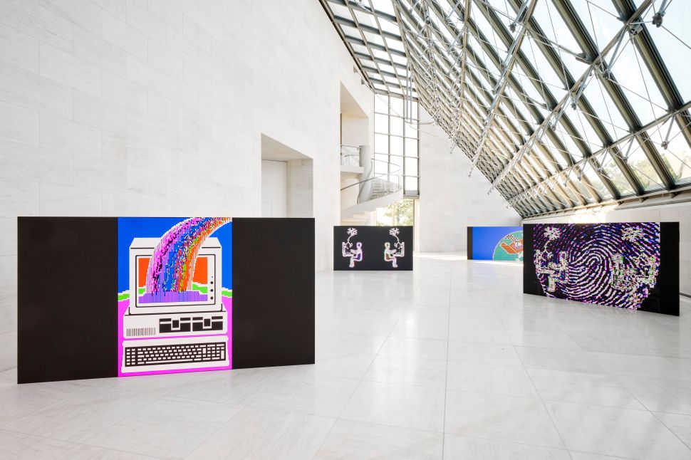 In a spacious, light-filled gallery with large windows and high ceilings, multiple digital artworks are displayed on freestanding black panels. The artworks feature colorful, pixelated images, including a rainbow flowing out of a computer and abstract patterns, with natural light streaming in from the windows above.