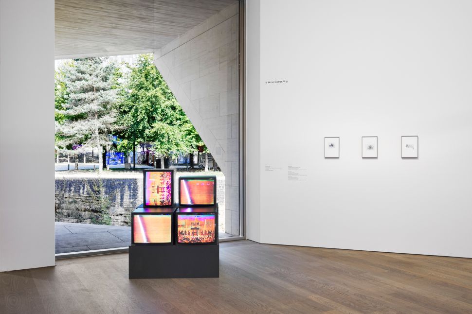 Inside a gallery space, a small stack of television monitors displaying bright, distorted images stands near a large window that looks out onto a green, tree-lined outdoor scene. On the adjacent wall are three framed works in a row, each with accompanying labels.