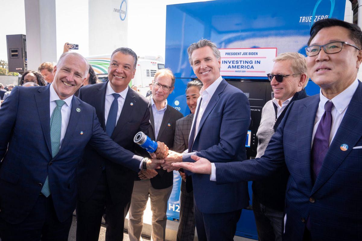 Men in suits stand around California governor Gavin Newsom, who is holding something in his hand that everyone is pointing to.