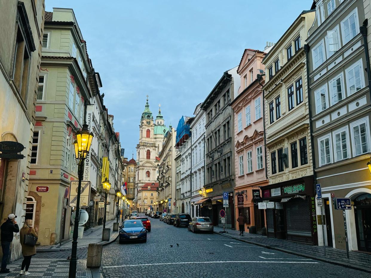 A few people walking down a quiet street in Prague at twilight.
