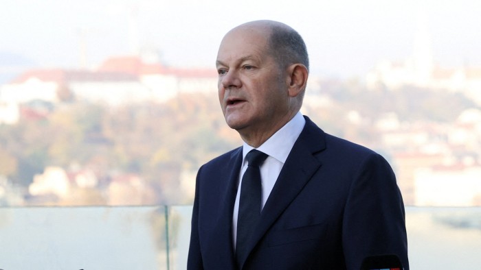 Olaf Scholz speaks to the media, standing outdoors with a blurred cityscape in the background.