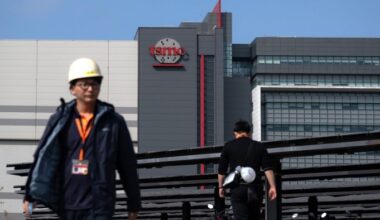 The Taiwan Semiconductor Manufacturing Company logo is prominently displayed on a building at the Hsinchu Science Park in Taiwan. In the foreground, two people wearing work attire and hard hats are walking