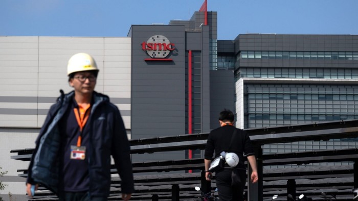 The Taiwan Semiconductor Manufacturing Company logo is prominently displayed on a building at the Hsinchu Science Park in Taiwan. In the foreground, two people wearing work attire and hard hats are walking