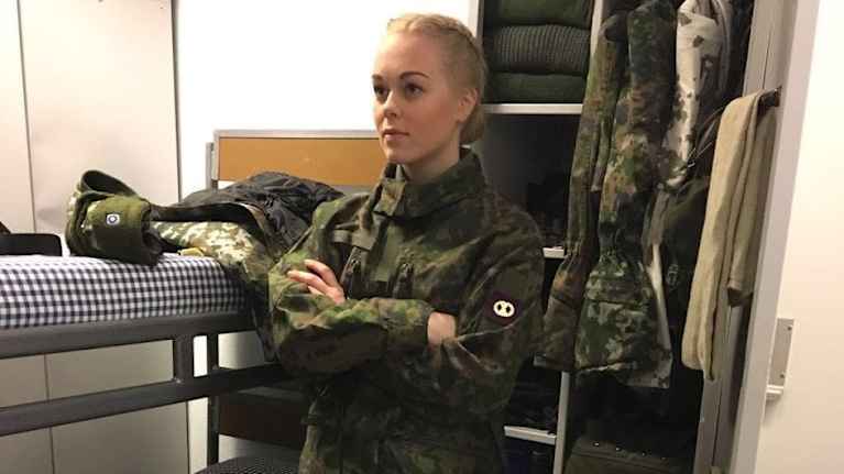 Parikymppinen Krista Liesniemi seisoo Säkylän varuskunnan tuvassa armeijan maastopuku yllään. Hänen takanaan näkyvät pedattu punkka ja avoinainen varustekaappi.