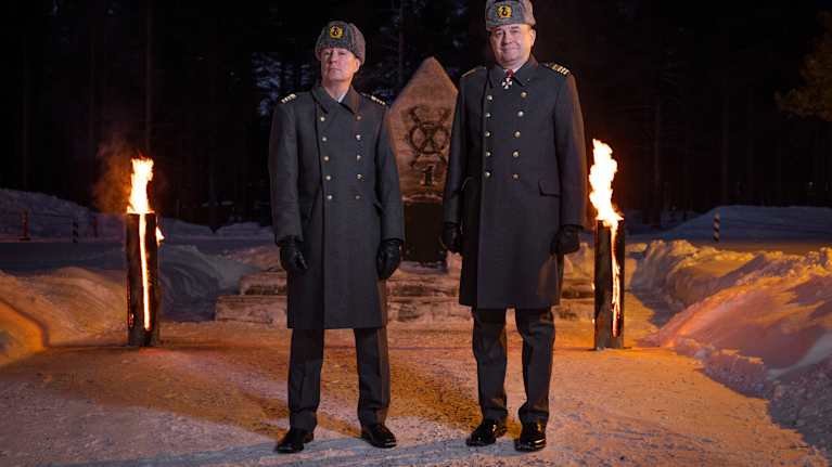 Kaksi everstiä takit päällään seisoo patsaan ja soihtujen edessä.