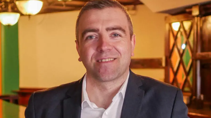 A man with short brown hair in a black blazer and white shirt looks directly at camera while vaguely smiling.