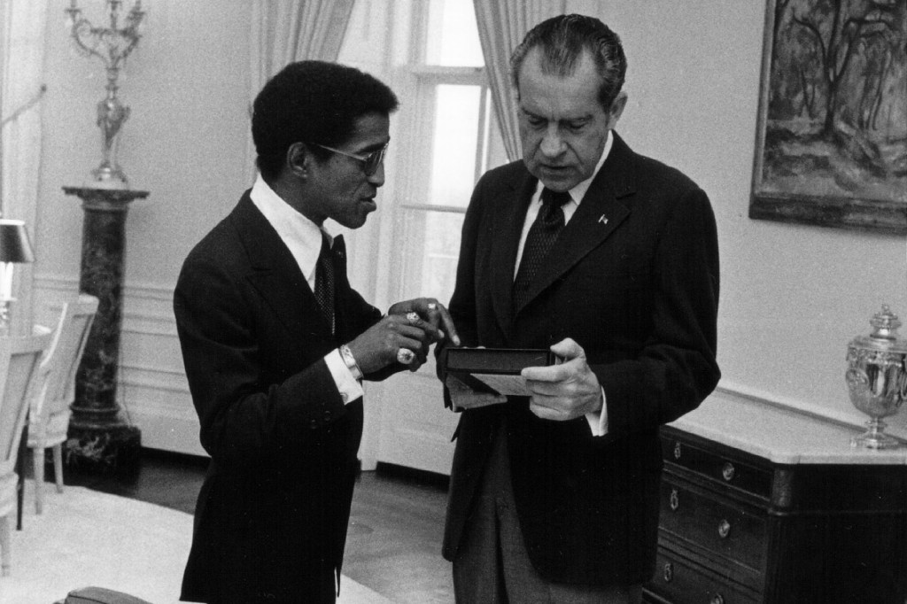 Richard Nixon in a suit and tie looking at Sammy Davis Sr., as narrated by comedians on a podcast.