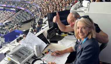 Two people in the press box at a stadium - one is giving the camera the thumbs up
