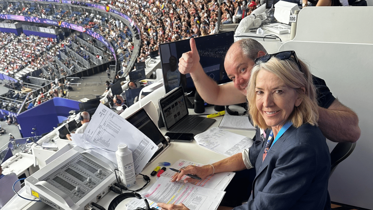 Two people in the press box at a stadium - one is giving the camera the thumbs up