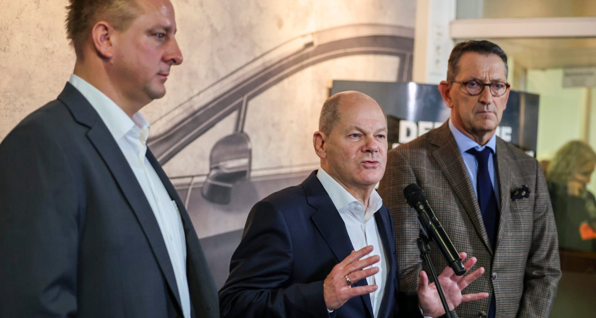 Bundeskanzler Olaf Scholz (SPD) steht zwischen dem Betriebsratschef von Ford Deutschland, Benjamin Gruschka (links), und dem Geschäftsführer der Ford Werke, Marcus Wassenberg (rechts).
