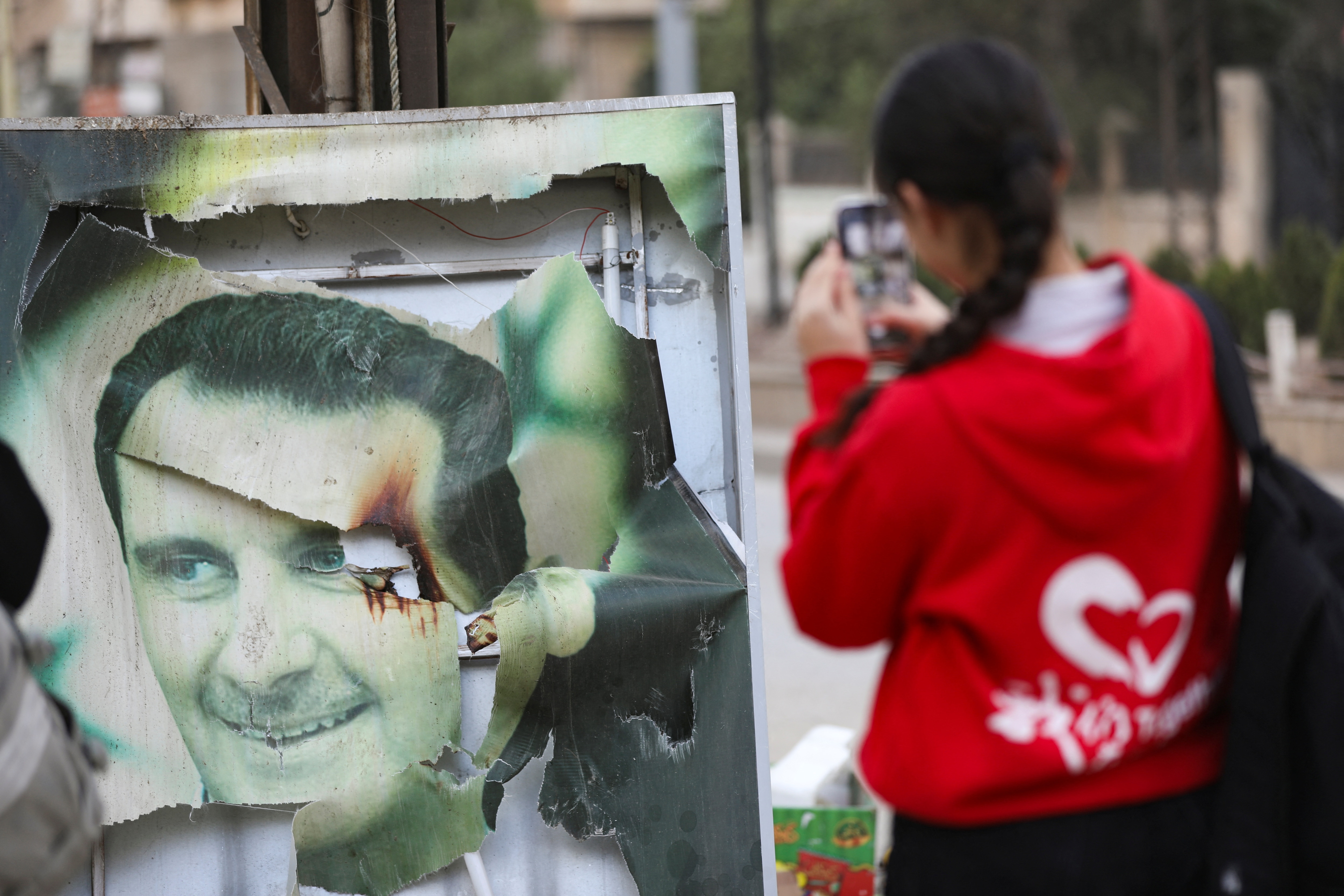 A woman uses her mobile phone near a damaged picture of Syrian President Bashar al-Assad as people celebrate, after Syrian rebels announced that they have ousted President Bashar al-Assad, in Qamishli, Syria December 8, 2024. REUTERS/Orhan Qereman