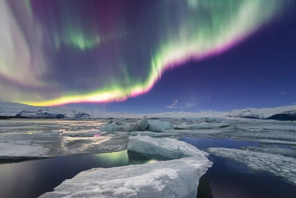 The northern lights shimmer in green and pink hues over Jökulsárlón Glacier Lagoon, with icebergs floating in the tranquil water and snow-capped mountains in the distance