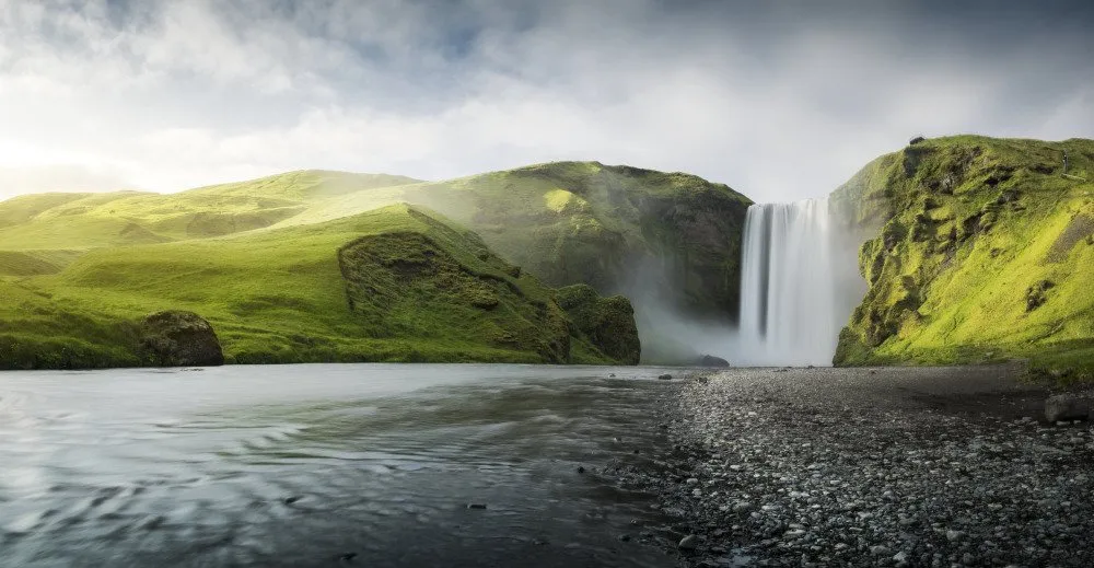 The Skógafoss waterfall cascades down a lush green cliffside, with a tranquil river flowing in the foreground 