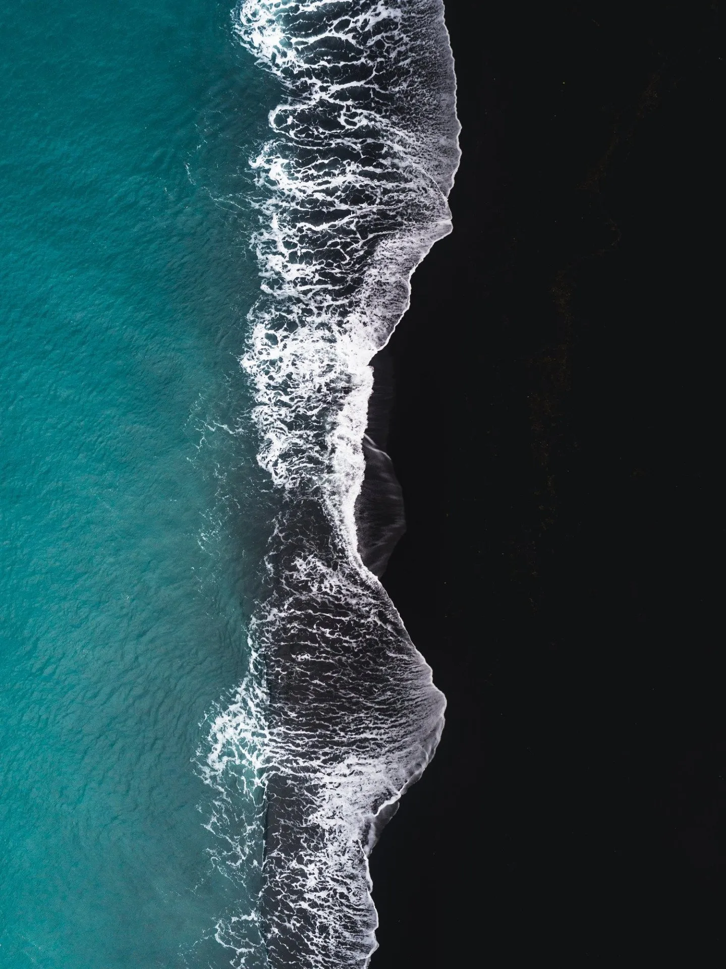 Aerial view of ocean waves crashing against a black sand beach, showcasing Iceland's striking natural contrast