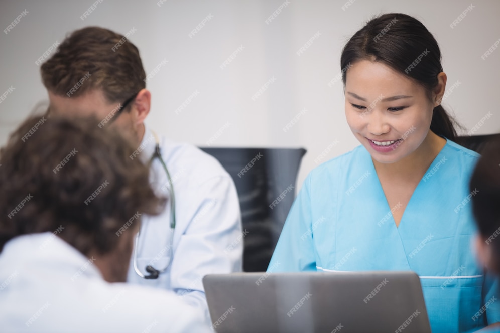 A smiling healthcare worker in blue scrubs works on a laptop, surrounded by colleagues in a professional setting.