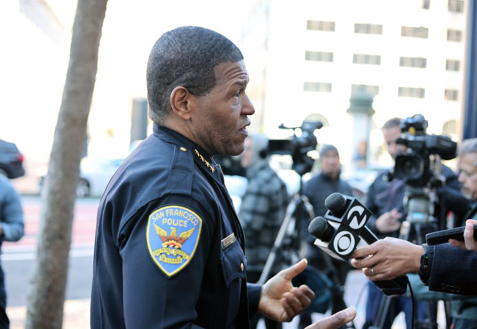 A police officer in uniform speaks to a group of reporters with microphones and cameras in an outdoor setting.