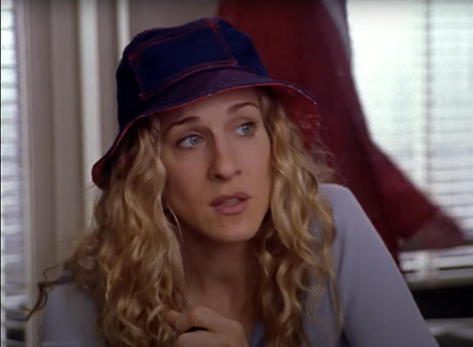 From "Sex and the City": Sarah Jessica Parker wearing a casual outfit and a bucket hat, sitting indoors and looking to the side with an unsure expression