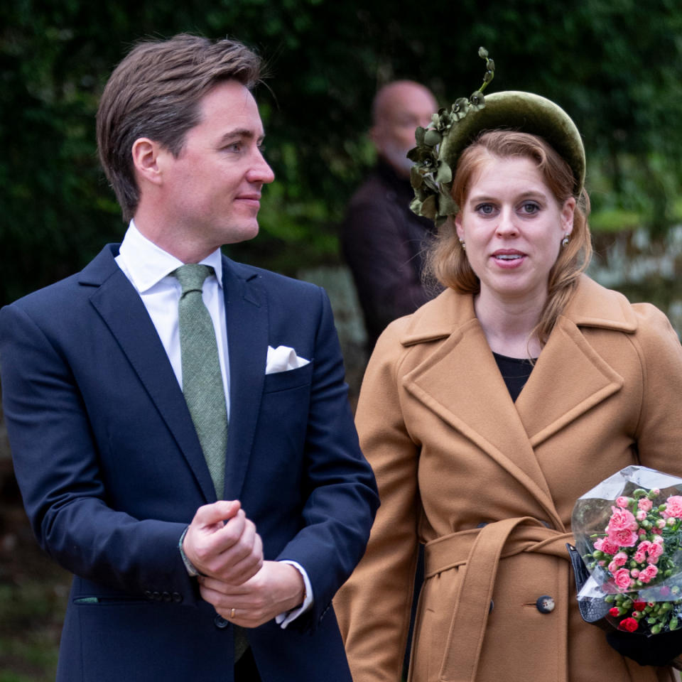  Princess Beatrice wearing a camel wrap coat and green headband holding flowers standing next to Edoardo Mapelli Mozzi who is wearing a navy suit and green tie. 