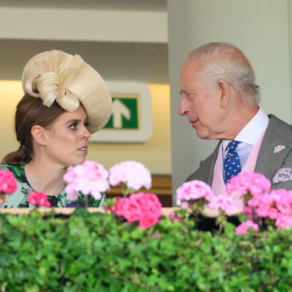 Princess Beatrice wearing a straw hat and green dress speaking with King Charles in a gray suit at Royal Ascot with pink flowers in the foreground.