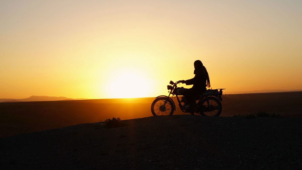 Cutting_Through_Rocks_اوزاک یوللار-Still3 | Filmmaker Magazine A silhouetted Iranian woman is riding a motorcycle against the sunset.