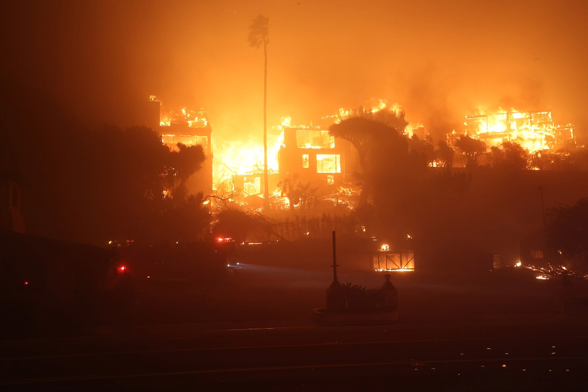 The Palisades Fire glows orange as it burns through at least three structures at night with trees in the foreground.
