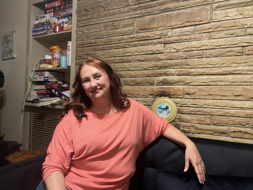 A woman with red hair rests with her arm on a sofa. She wears a salmon-colored blouse.
