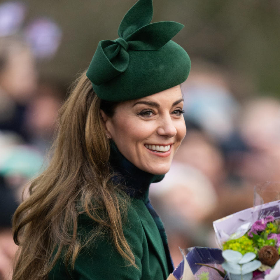 Kate Middleton wearing a green hat and coat smiling and holding flowers with a crowd of people blurred behind her.